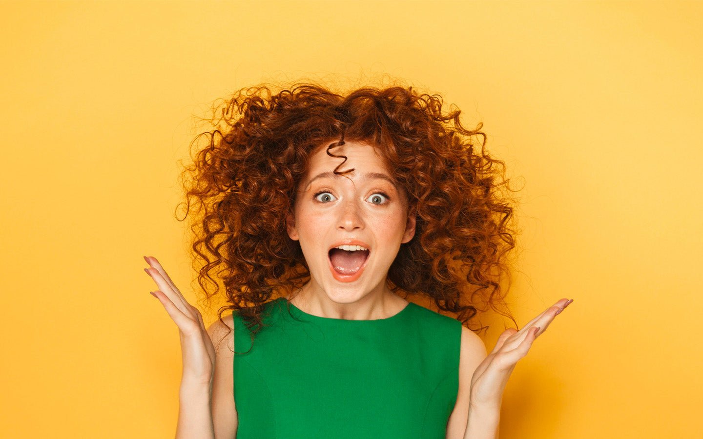 Surprised woman with red curly hair and green top, hands raised, on yellow background