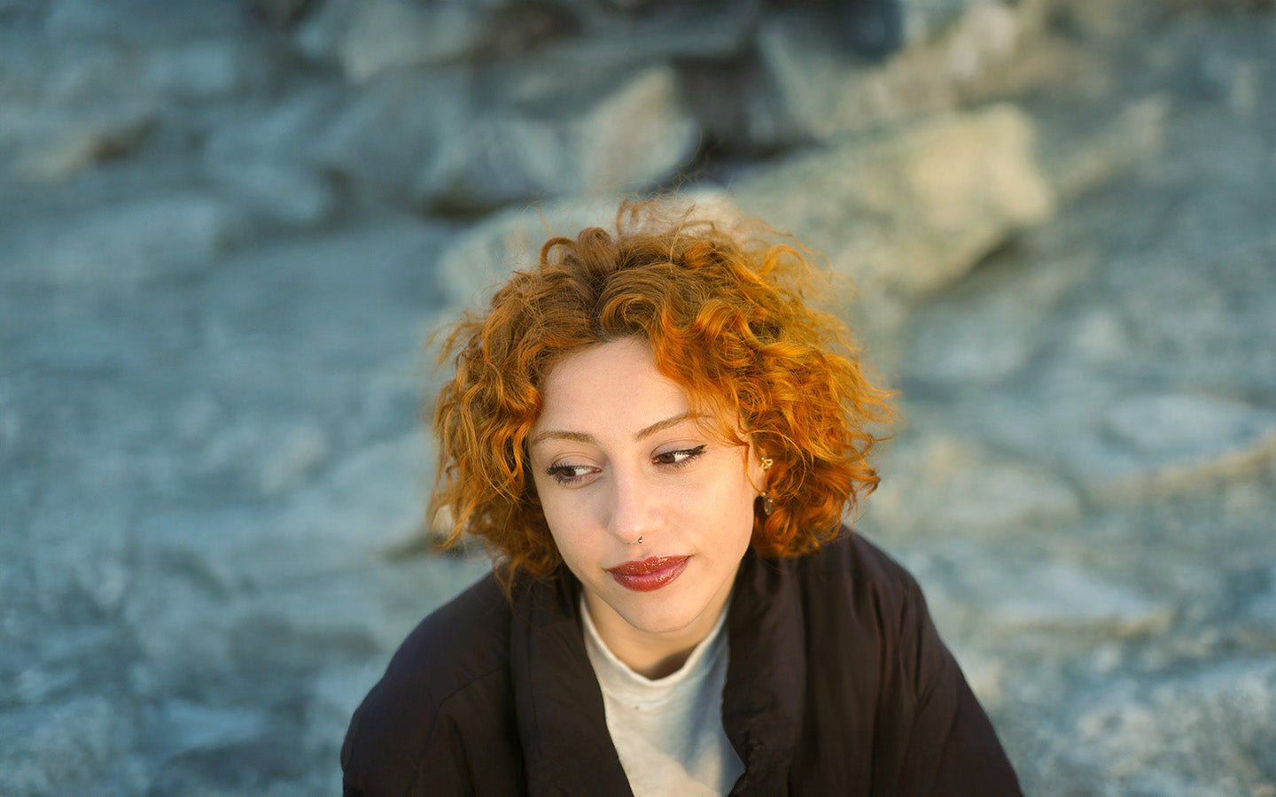 Woman with red curly hair, nose ring, and dark jacket looking down