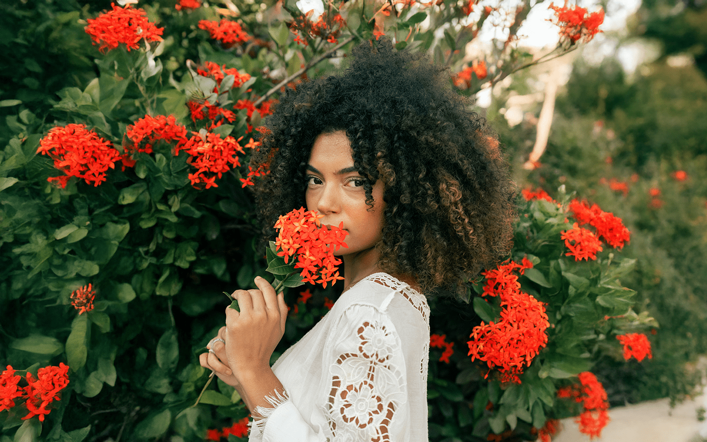 Woman with curly hair in a white lace top smelling red flowers outdoors