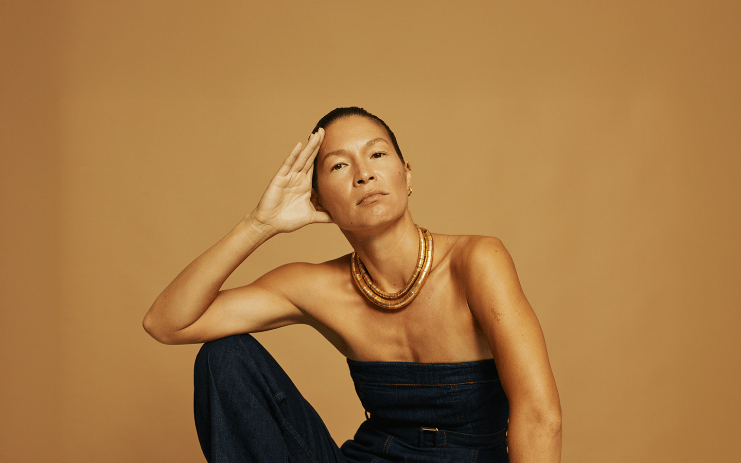 Woman in denim tube top and gold necklace, hand resting on her head