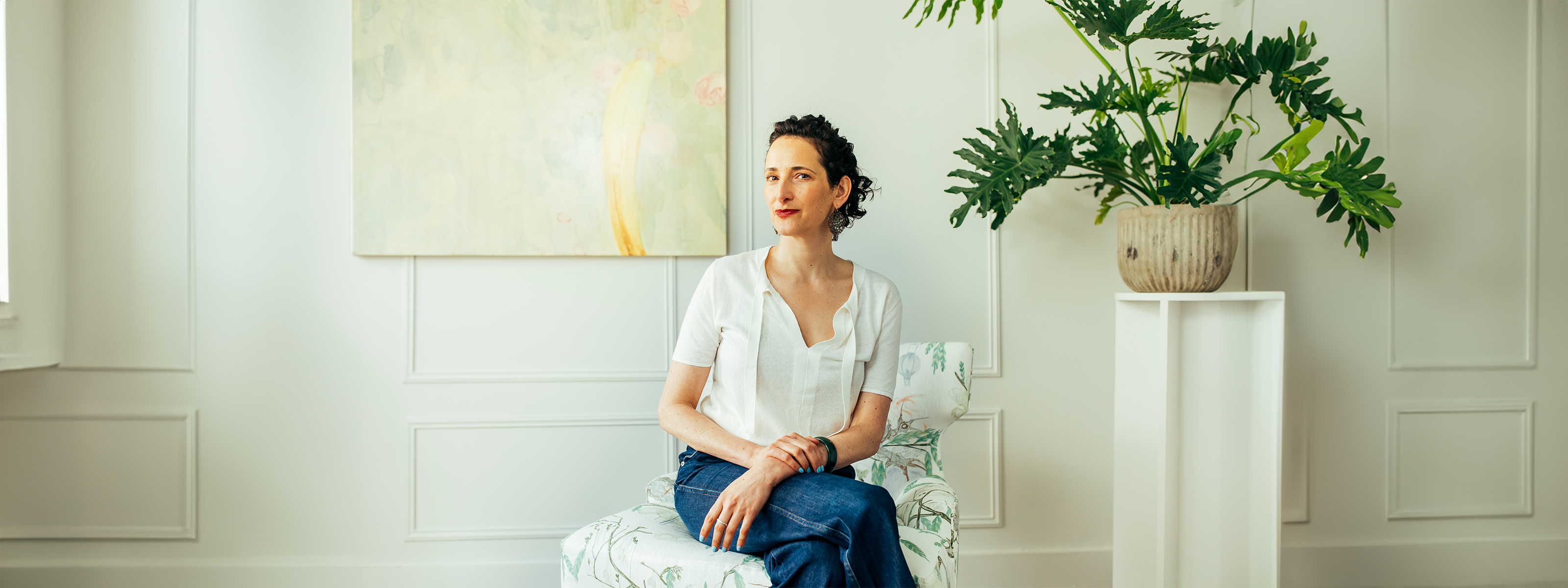 Beautiful woman seated blue jeans cream blouse plant on stand and painting in background