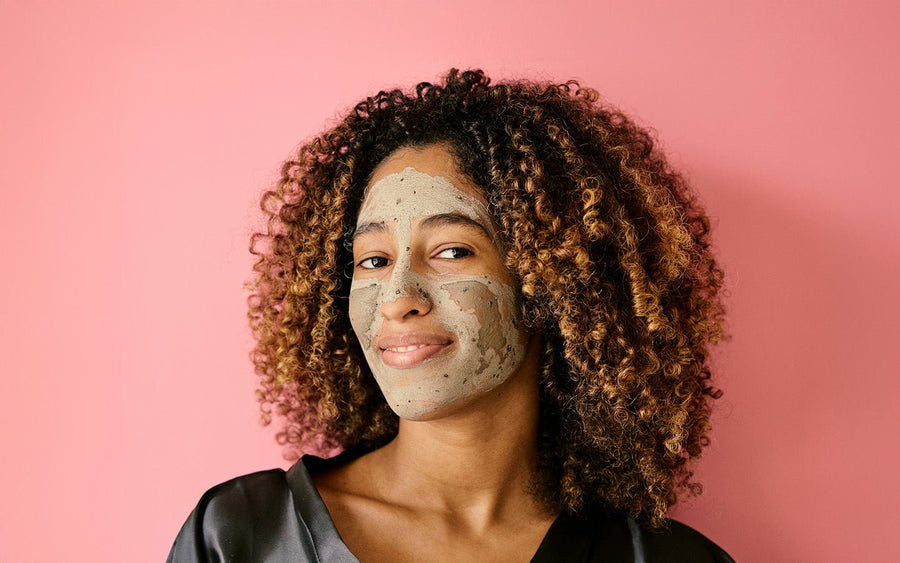 Woman with curly hair and a grey face mask smiles against a pink background