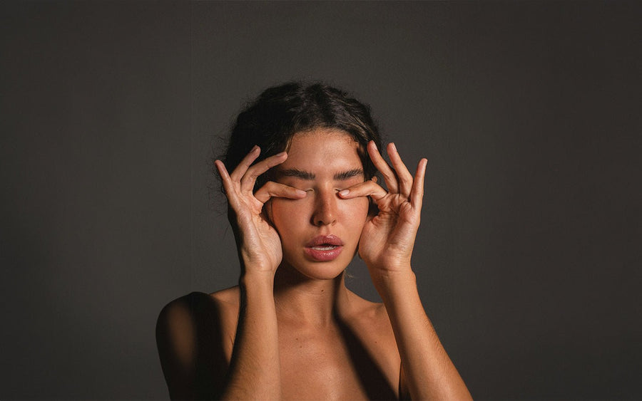 Woman with closed eyes, fingers gently pressing on eyelids, against a dark gray background