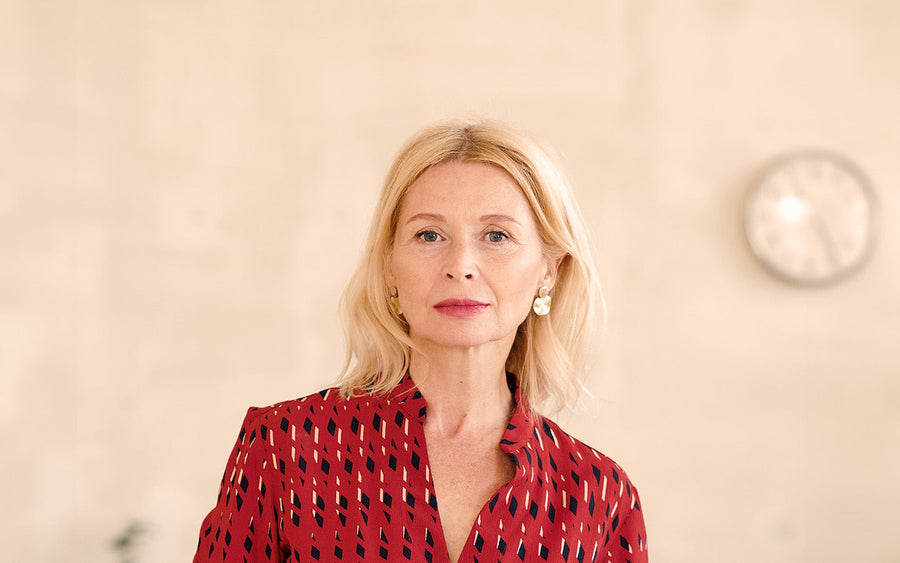 Blonde woman in red patterned shirt with gold earrings, light background, blurred clock