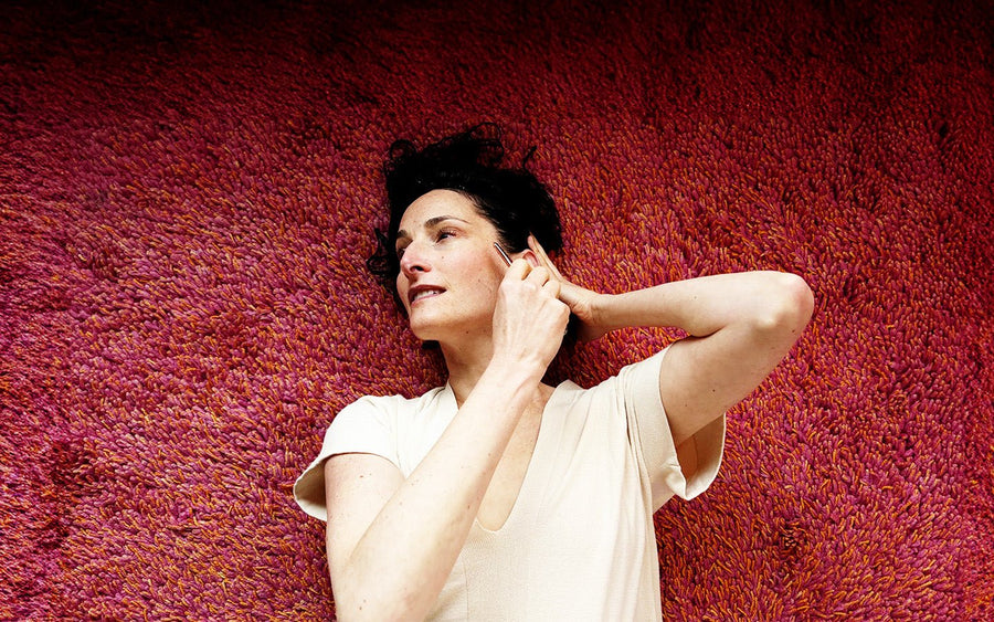 Woman in cream shirt lies on a red shag rug, using a gua sha tool on her face