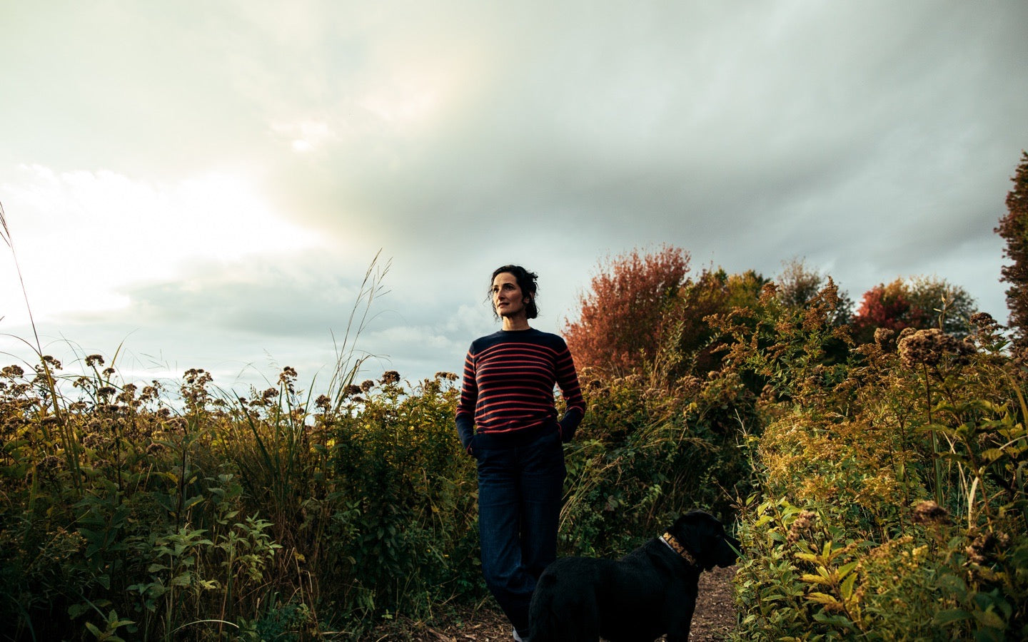 Dr. Catherine standing with a black dog among autumn plants beneath a cloudy sky.
