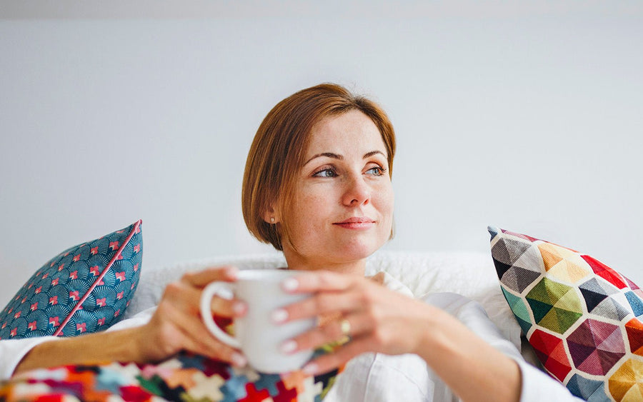 Woman with short brown hair holding a white mug, sitting on a white couch with colorful pillows