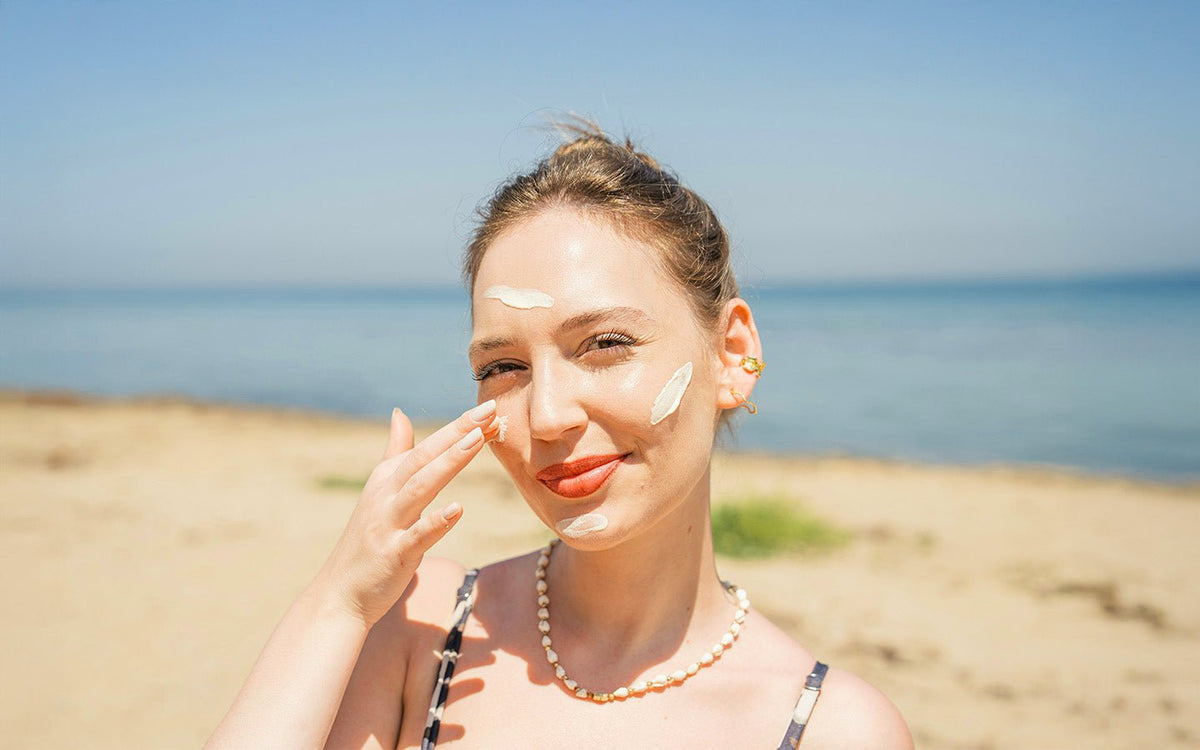 Woman on beach applying white sunscreen to her face, wearing a pearl necklace