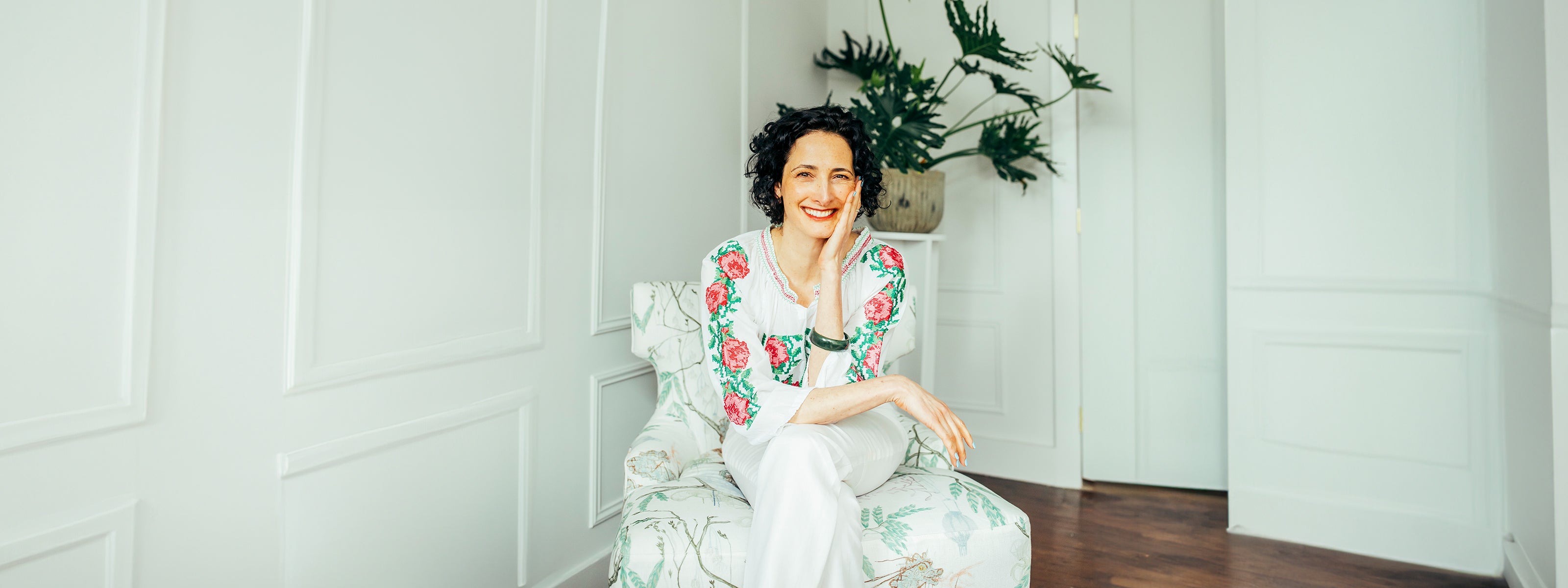 Smiling woman red lipstick seated with floral blouse white pants